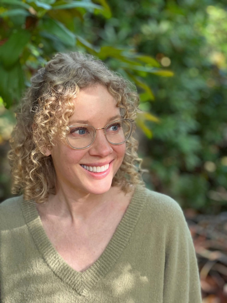 Shannon Tully, white woman with blond curly hair and wire rim glasses, smiling outside in nature.